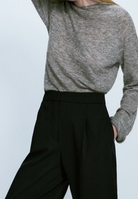 Personne portant un pull léger à manches longues gris clair rentré dans un pantalon noir taille haute, une main dans la poche et le bras levé.