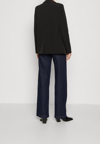 Černý blazer se strukturovaným střihem a ostrými klopami, zkombinovaný s tmavomodrými širokými džíny a černými botami na podpatku. Hladká textilie.