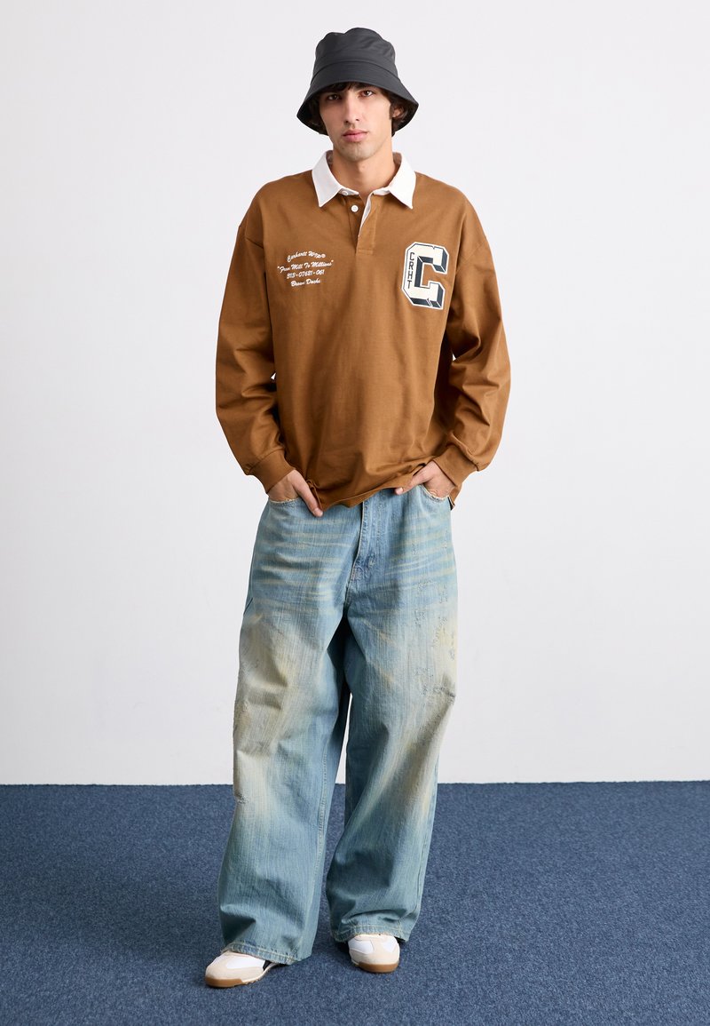 Polo shirt marron à manches longues avec col blanc et graphique sur la poitrine, associé à un jean large bleu clair avec une finition déchirée. Baskets blanches.