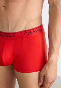Boxer briefs rojos de Calvin Klein con una cintura de logo negro. La tela parece suave y ajustada, mostrando un color sólido sin patrones.