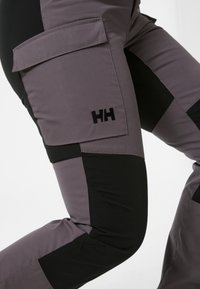 Pantalones cargo gris y negro con un corte ajustado, que cuentan con un gran bolsillo lateral y un logo bordado en el muslo. Textura de tela suave.
