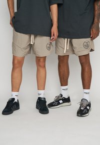 Pantalones cortos beige con un logo en la pierna izquierda, cintura con cordón y una textura ligera. Los lleva puestos con zapatillas negras y calcetas blancas.