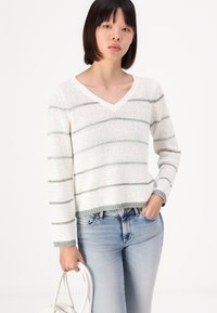 Hvit strikkegenser med grønne horisontale striper, v-hals-design og ribbet kant. Kombinert med lyseblå jeans og holder en hvit veske.