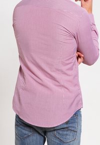 Muž v dlouhé růžové kostkované košili a modrých džínách, stojící s založenýma rukama, otočený zády k fotoaparátu.