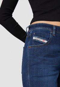 Jeans de ganga azul escuro com um design slim-fit. Apresenta costura laranja, um rótulo no bolso de trás e um estilo clássico de cinco bolsos.