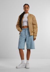 Hnedá bomberová bunda, biely crop top, svetlomodré džínsy šortky, biele tenisky, v kombinácii s bielymi ponožkami. Model stojí pred sivým pozadím.