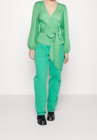 Zelená blůza s volnými rukávy, texturovaný materiál a vázání v pase, v kombinaci se zelenými cargo kalhotami. Černé kotníkové boty.
