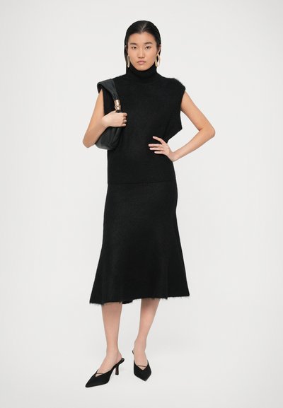 Haut sans manches en mélange de laine noire avec col roulé, associé à une jupe évasée assortie, offrant une texture douce. Complété par un sac noir et des talons.