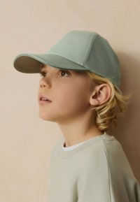 Jeune enfant aux cheveux blonds bouclés portant une casquette et un sweat-shirt vert pâle, regardant vers le haut sur un fond de mur beige.