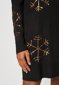 Melna adīta džemperis ar zelta sekmēm sniega более dizainu, ribotām piedurknēm un apakšmalu, kā arī mīkstu tekstūru.