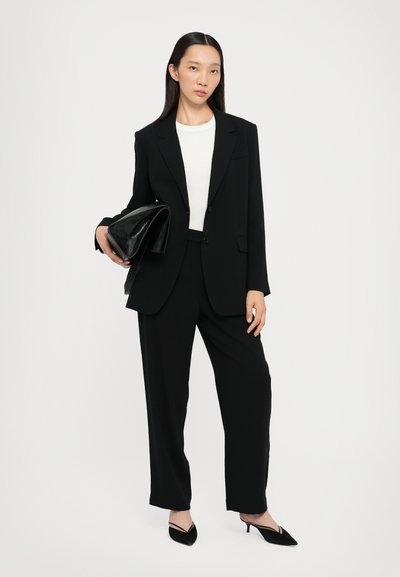 Schwarzer maßgeschneiderter Anzug mit strukturiertem Blazer und lockeren Hosen. Das Modell hält eine schwarze lackierte Clutch. Weißes Oberteil darunter, schlanke Silhouette.