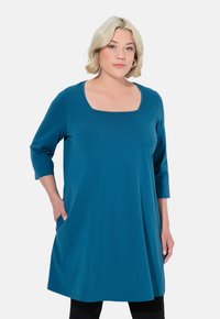 Tunika-Kleid in Türkis aus weichem Stoff, mit quadratischem Ausschnitt und Dreiviertelärmeln, seitlichen Taschen und einem lockeren Schnitt.