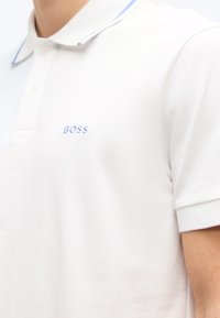 Κοντινό πλάνο ενός ατόμου που φοράει λευκό πόλο μπλουζάκι με γιακά με μπλε περίγραμμα και μικρό κεντημένο λογότυπο "BOSS" στο στήθος.