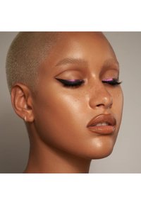 Nærbilde av en modell med kort blondt hår, iført kraftig svart eyeliner med rosa aksenter, jevn brun leppestift, og en glowy hudtone.