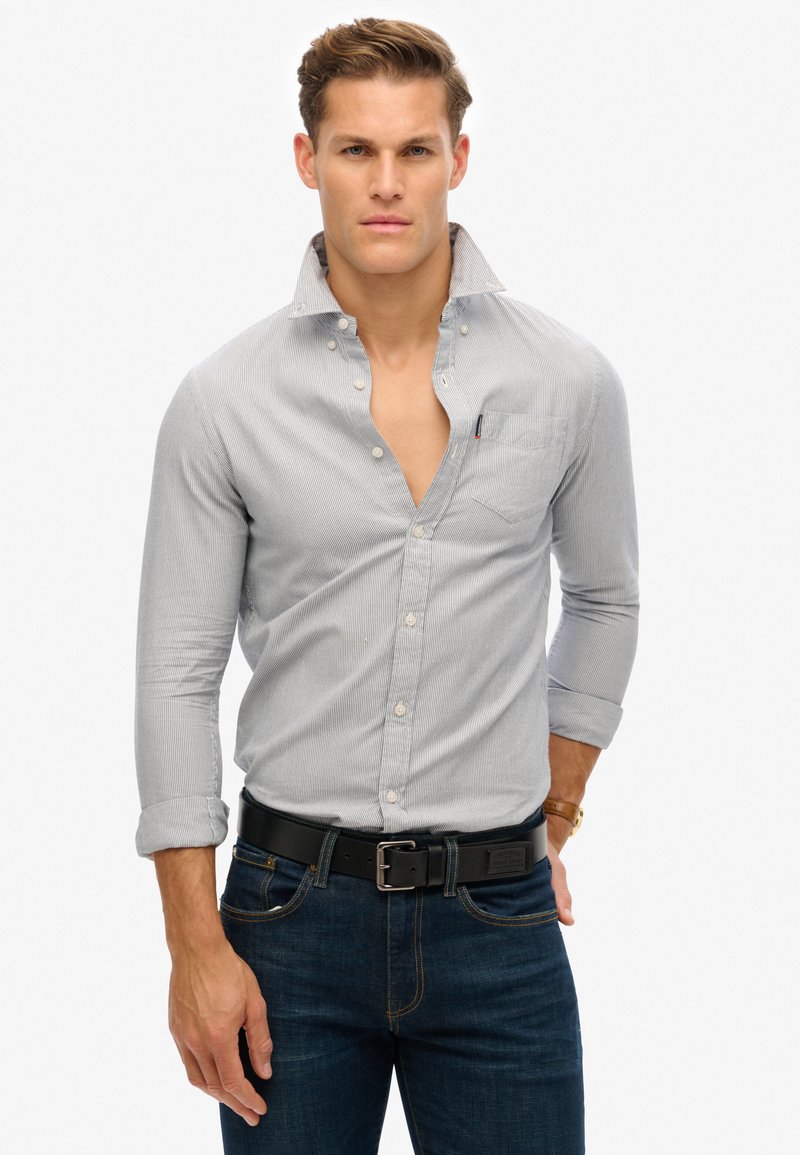 Camicia button-up con un motivo a righe blu chiaro e bianche, caratterizzata da una tasca sul petto e colletto aperto, abbinata a jeans blu scuro e una cintura nera.