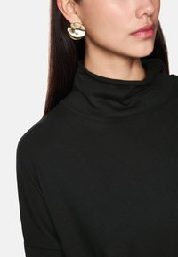 Schwarzes Rollkragenoberteil aus weichem Stoff mit hohem Kragen. Goldene kreisförmige Ohrringe reflektieren das Licht und setzen einen Kontrast zum Outfit.