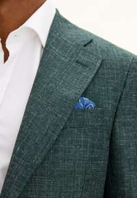 Vihreä teksturoitu bleiseri, jossa on viistot läpät, istuva siluetti ja sininen taskuliina rintataskussa valkoisen paidan päällä.