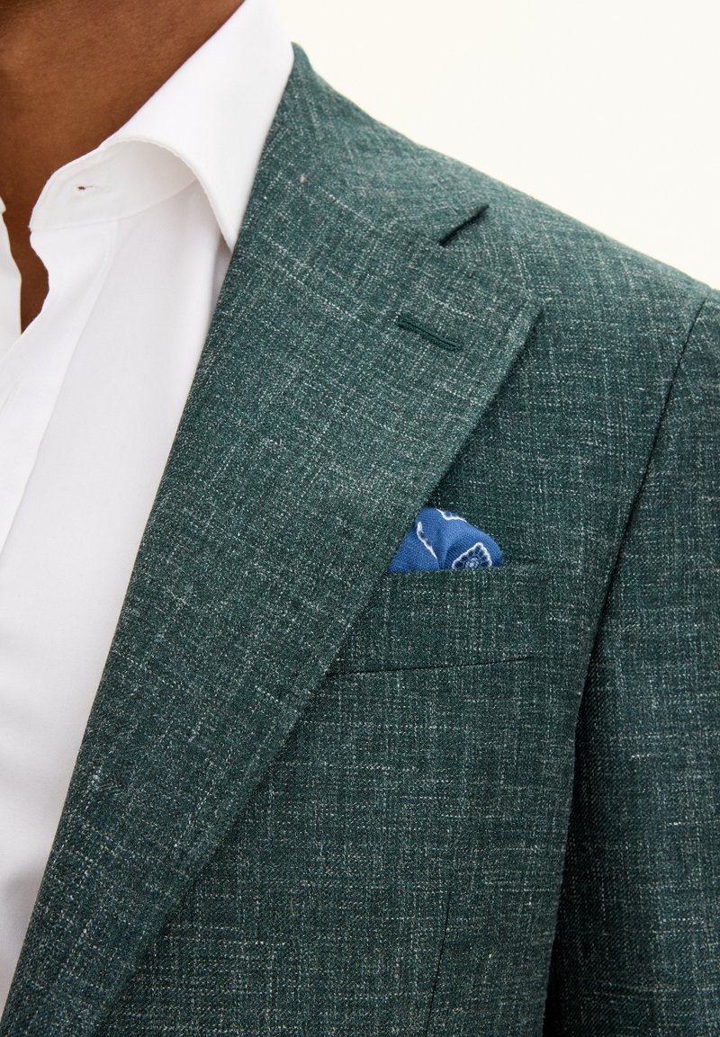 Vihreä teksturoitu bleiseri, jossa on viistot läpät, istuva siluetti ja sininen taskuliina rintataskussa valkoisen paidan päällä.