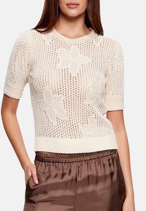 Donna che indossa un top crema all'uncinetto a maniche corte con motivi floreali e pantaloni di raso marroni a vita alta, mano in tasca.