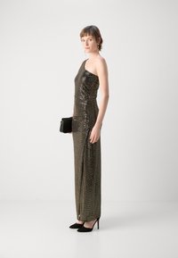 Robe de soirée métallique à une épaule avec une silhouette ajustée. Dotée d'embellissements scintillants et d'une fente sur le côté. Associée à un petit sac à main noir et des talons.
