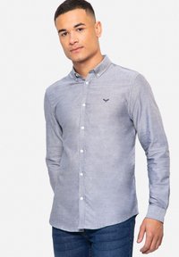 Blauwe blouse in een lichte stof. Met een kraag, witte knopen en een embleem op de borst. Gecombineerd met donkere jeans.