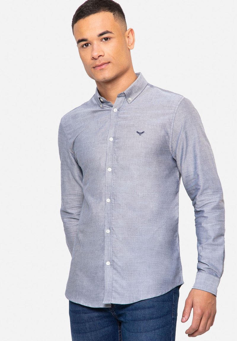 Blauwe blouse in een lichte stof. Met een kraag, witte knopen en een embleem op de borst. Gecombineerd met donkere jeans.