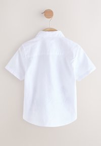 Chemise blanche à manches courtes avec col boutonné, empiècement dos, et ourlet arrondi, présentée sur un cintre en bois.