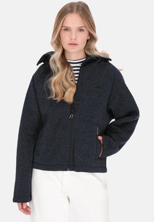 Junge Frau mit langen blonden Haaren, die eine dunkle Jacke mit Reißverschluss über einem gestreiften Shirt und weißen Hosen trägt, steht mit einer Hand in der Tasche.