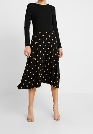 Nainen pukeutunut mustaan pitkähihnaiseen toppiin ja mustaan midi-hameeseen, jossa on beigejä pilkkuja, yhdistettynä beigeihin avokkaisiin korollisiin sandaaleihin.