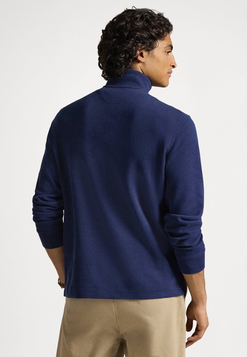 Man with curly hair wearing a navy blue turtleneck sweater and beige pants facing sideways against a plain white background.