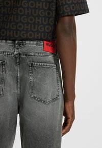 Maglietta nera a maniche corte con logo "HUGO" ripetuto, abbinata a jeans in denim grigio con cintura brandizzata rossa e tasche posteriori.