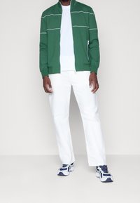 Giacca verde con zip e strisce bianche, t-shirt bianca, pantaloni bianchi e scarpe da ginnastica blu. Tessuto liscio, vestibilità casual e design moderno.