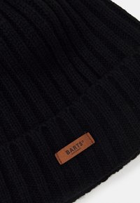 Svart stickad mössa med ribbad textur. Har en brun läderetikett präglad med "BARTS AMSTERDAM" nära den nedre kanten.