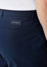 Pantalones azul marino con una tela texturizada y una etiqueta que dice "HACKETT". Bolsillos laterales visibles, diseño ajustado resaltado.