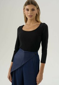 Zwarte lange mouwen top met een ronde hals, gecombineerd met een blauwe wikkelrok met een diagonale plooi en knoopdetail.