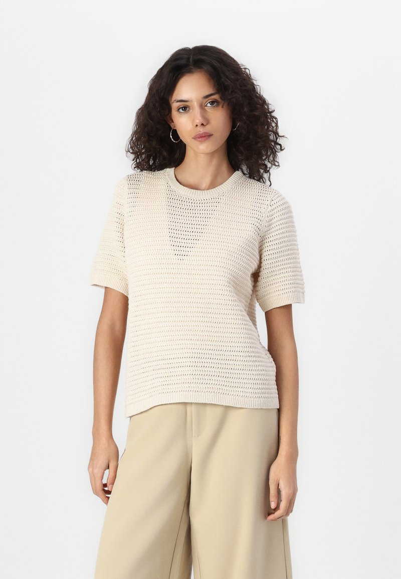 Beige gebreid sweater met korte mouwen, ronde halslijn en een gestructureerd patroon. Gepaard met lichtbruine wijde broek.