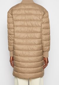 Béžový prošívaný kabát s vodorovným prošíváním, dlouhými rukávy a žebrovanými manžetami. Hladká textura a délka sahající ke kyčlím.