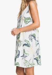 Robe ample sans manches en tissu blanc avec motifs de feuilles tropicales vertes et fleurs. Encolure ronde et découpes latérales pour une meilleure aération.