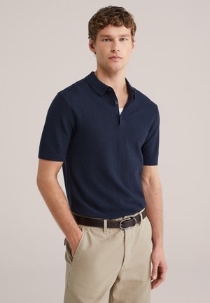 Jonge man die een marineblauw poloshirt met korte mouwen, beige broek en een donkerbruine riem draagt, staand met één hand in zijn zak.