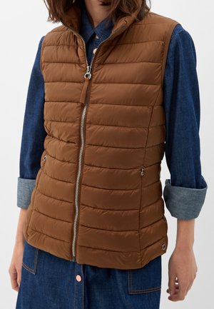 Vrouw draagt een bruine gewatteerde mouwloze bodywarmer met ritssluiting over een blauwe denim blouse met lange mouwen en opgerolde manchetknopen.