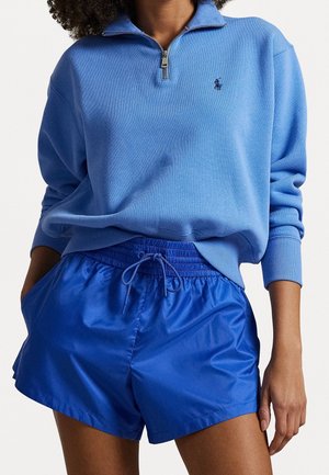 Osoba ubrana w jasnoniebieską długorękawową bluzę z zamkiem do ćwiartki i błyszczące niebieskie szorty z elastycznym pasem i sznurkiem, stojąca z rękami w kieszeniach.