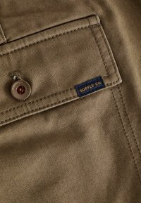 Khaki stof met een gladde textuur, met een close-up van een zak, een metalen knop en een blauwe label met de tekst "SUPPLY CO."