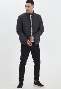 Grå puffjacka med hög krage, med en quiltad design och framdragkedja. Bärs över en vit skjorta med mönstrade detaljer och svarta byxor.