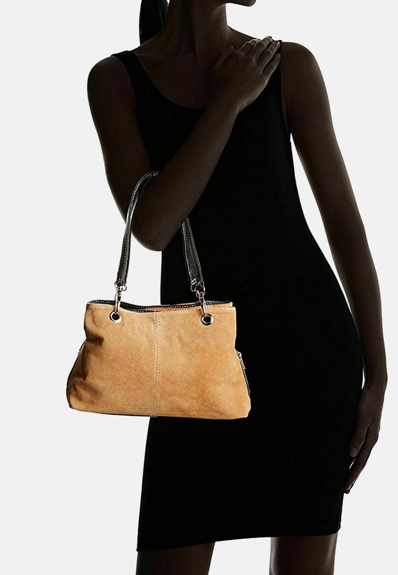 Femme en robe noire sans manches tenant un sac à main en daim beige avec des anses en cuir noir sur un fond uni.
