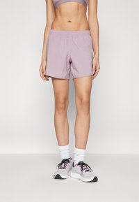 Shorts de sport lilas en matériau léger, avec une taille élastique et une coupe décontractée. Portés avec des chaussettes blanches et des chaussures de sport.