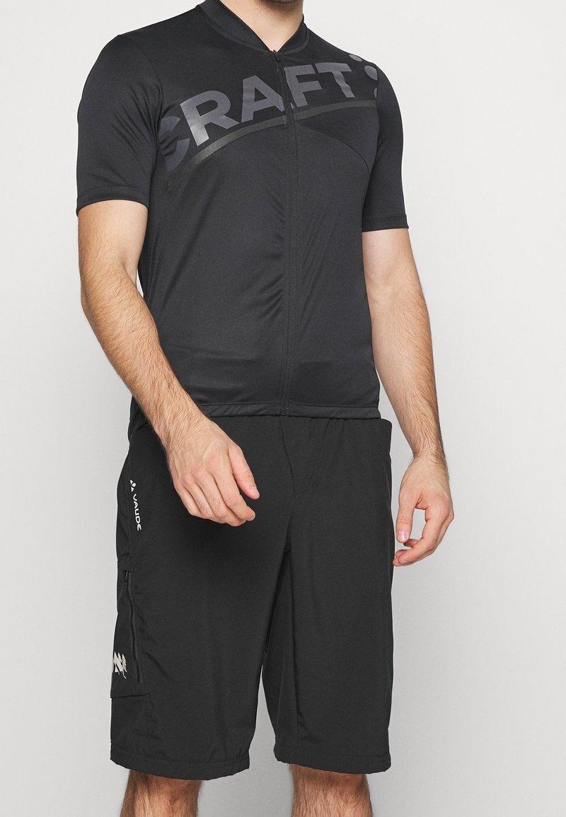 Maillot de cyclisme noir à manches courtes, avec une fermeture éclair sur le devant et une texture lisse. Associé à un short noir avec des poches zippées et des détails de marque.