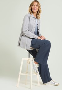 Jasnoszara kurtka zapinana na guziki z kołnierzykiem, zestawiona z granatowymi szerokimi spodniami i srebrnymi sneakersami, siedząca na białym stołku. Gładkie faktury.