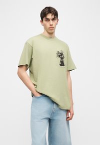 Νεαρός άνδρας με κοντά σκούρα μαλλιά, φορώντας πράσινο πουκάμισο χρώματος φασκόμηλου με μαύρο σχέδιο λουλουδιού και ανοιχτό μπλε τζιν, στέκεται με το ένα χέρι στην τσέπη.