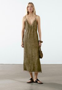 Olive green slip dress with a V-neck, adjustable straps, and gathered waist. Worn with black flat sandals and a small brown bag.