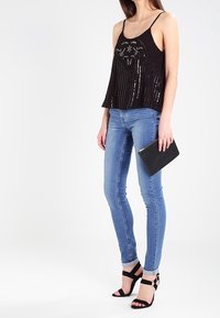 Débardeur en sequins noirs avec un motif en cœur, associé à un jean slim bleu clair et des talons aiguilles noirs à lanières, tenant une pochette noire.